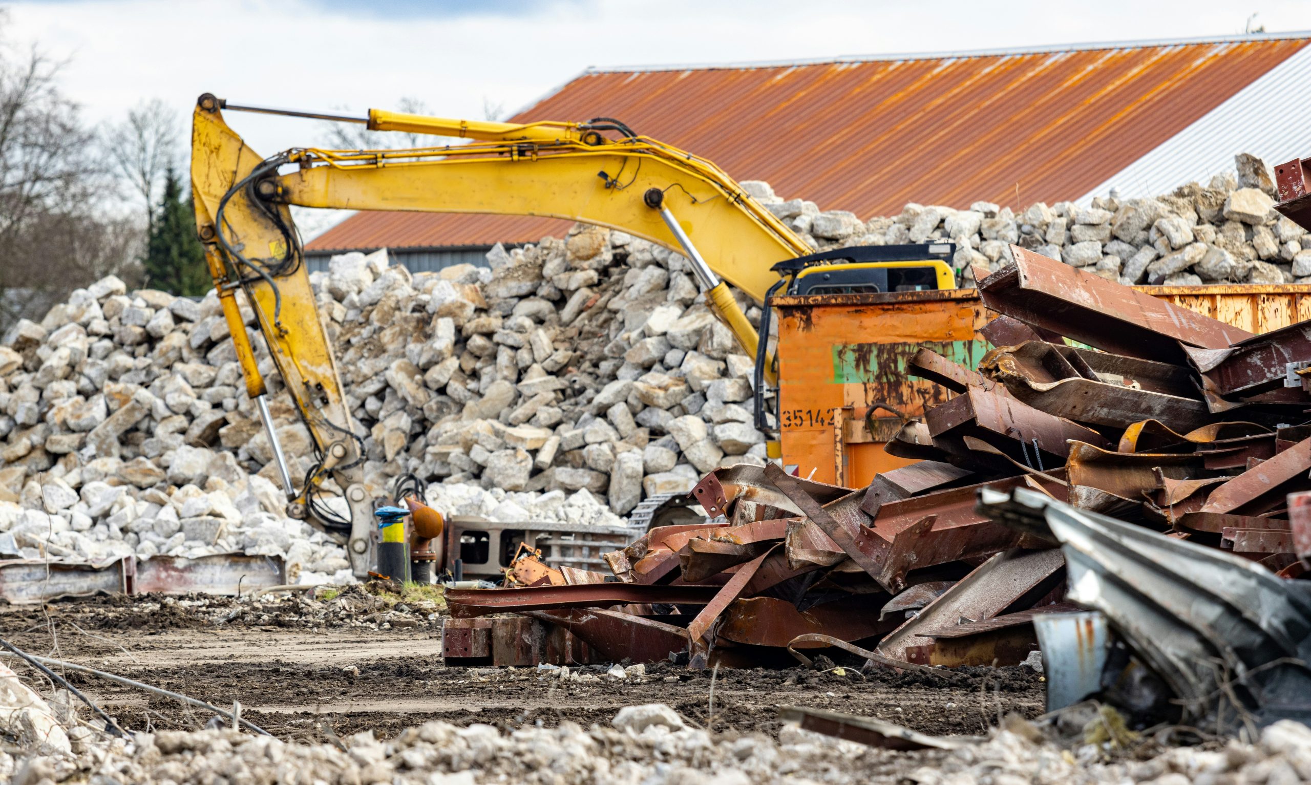 jobsite-waste-management-demolition-debris-excavator-bee Excavator with hydraulic breaker beside a steel skip and piles of twisted girders and concrete rubble on a demolition jobsite